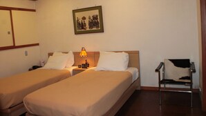 Quarto de hotel com duas camas de solteiro, uma mesa de cabeceira com abajur, uma cadeira e um quadro na parede.