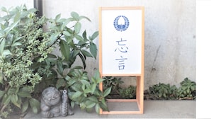 Uma placa de madeira com caracteres japoneses e um logotipo, colocada ao ar livre em frente a uma parede de concreto.