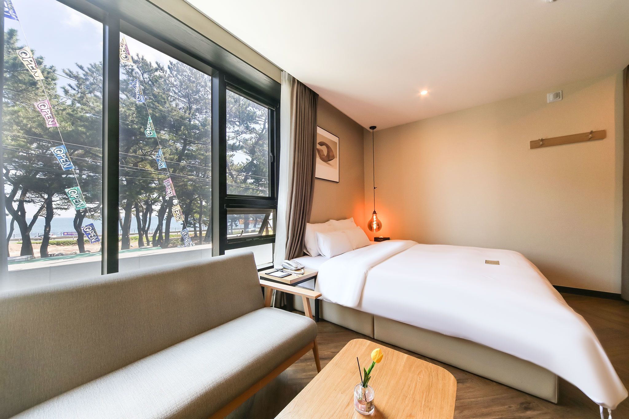 A hotel room with a bed, a sofa, a small table with a vase, a nightstand, and a window with curtains.