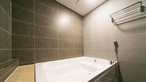 A modern bathroom with a large bathtub, dark tiles on the walls, and a shower area with a glass door. - Le Idea Hotel (Gyeongju)