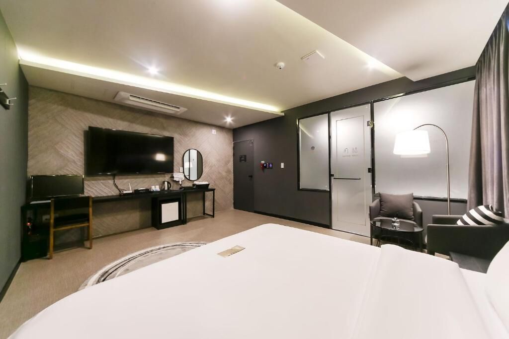 Moderni hotellihuone, jossa on suuri sänky, taulutelevisio, peilillä varustettu työpöytä, pieni pöytä sekä oleskelualue, jossa on tuoli ja lamppu.