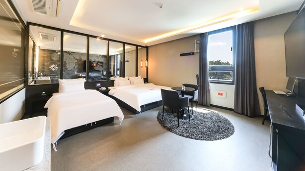 A hotel room with two beds, a desk, a chair, a round rug, and a large window with curtains. - Le Idea Hotel (Gyeongju)