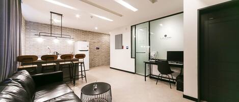 Une chambre d’hôtel moderne avec un canapé en cuir, une table basse ronde, un coin repas avec un mur en briques et un espace bureau séparé équipé d’un ordinateur.