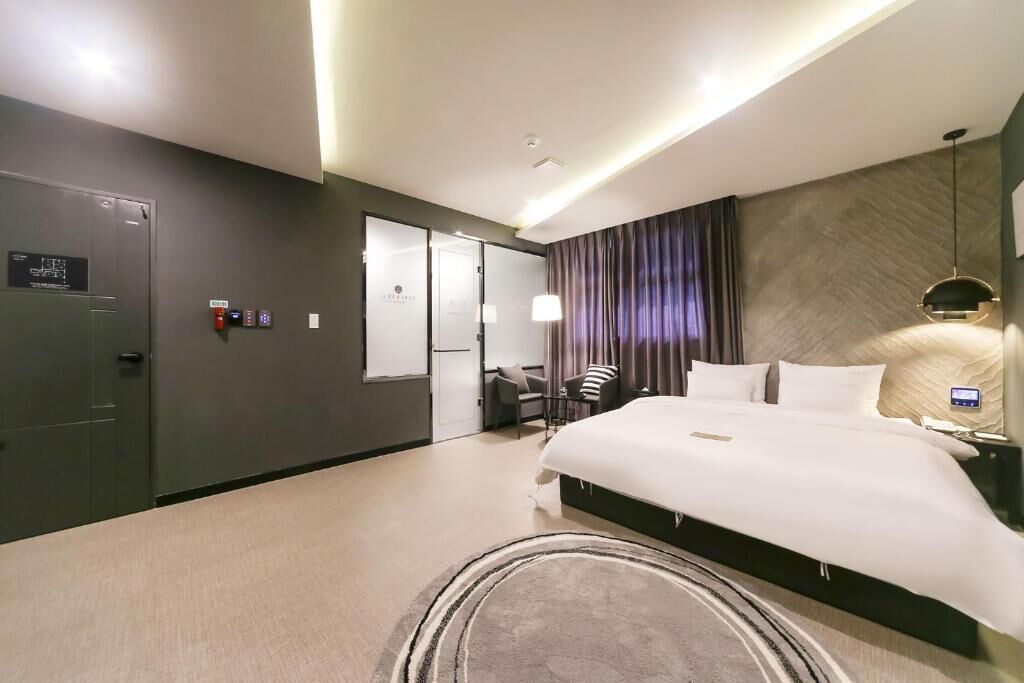 A modern hotel room with a large bed, a desk, and a chair. There is a wardrobe and a bathroom visible.