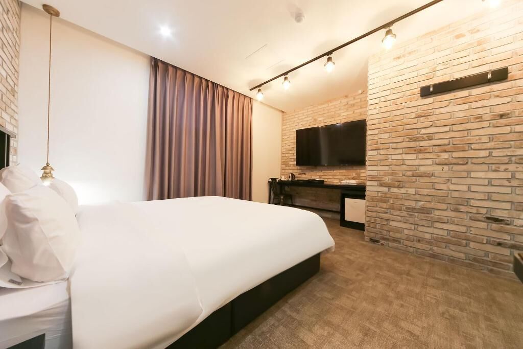 A modern hotel room with a large bed, a flat-screen TV mounted on a brick wall, a desk with a chair, and a curtain on the window.