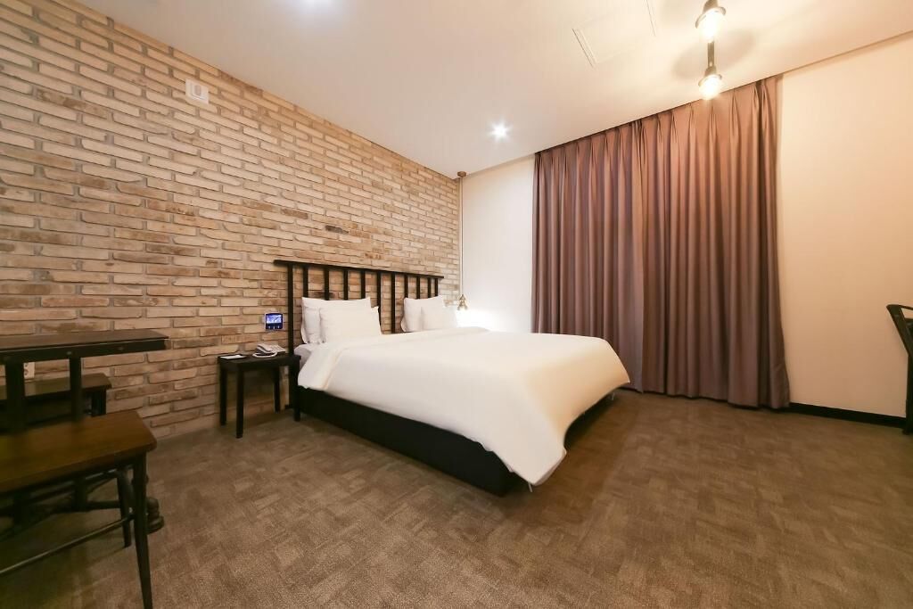 Une chambre d’hôtel avec un mur en briques, un lit, un bureau et une grande fenêtre avec des rideaux.