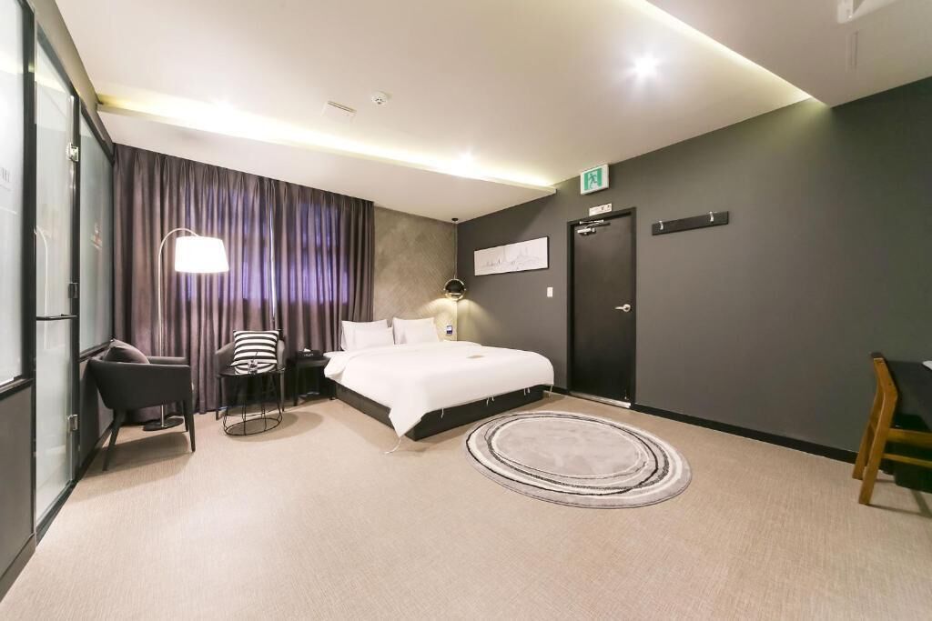 A modern hotel room with a bed, a desk, a chair, a lamp, and a door.