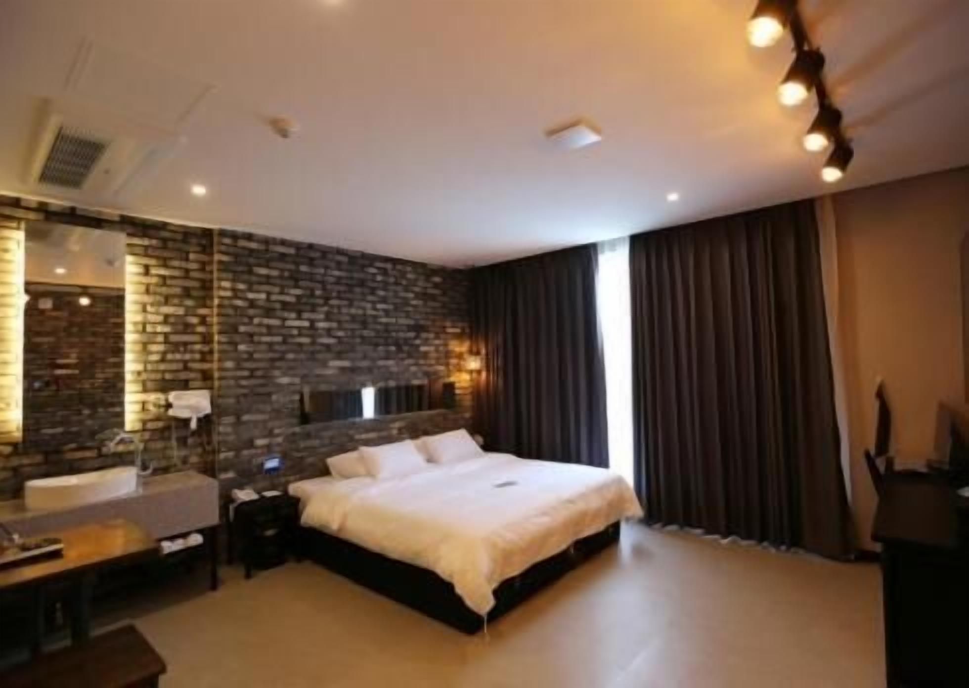 A modern hotel room with a large bed, a built-in desk, a mirror, and a stone accent wall.