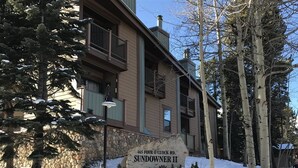 Un edificio de dos pisos con fachada de piedra, un letrero que dice "465 FOUR O'CLOCK RD. SUNDOWNER II", y alrededores cubiertos de nieve.