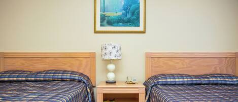 A hotel room with two beds, a nightstand with a lamp, a framed picture on the wall, and a small shelf with a book.