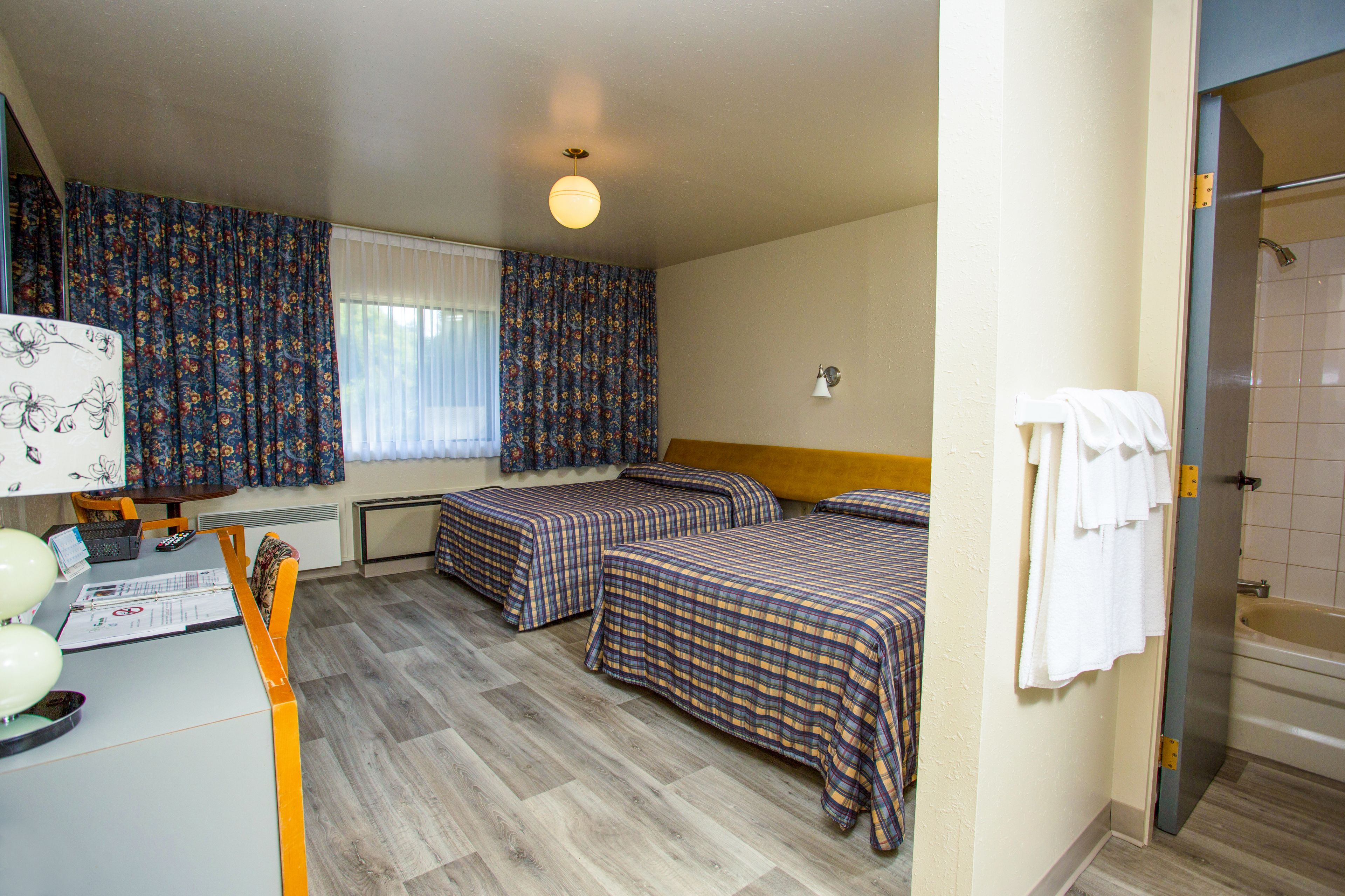 Une chambre d’hôtel avec deux lits, un bureau avec une lampe, une fenêtre avec des rideaux, une douche et une salle de bain avec une baignoire.
