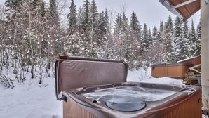 An outdoor hot tub with a wooden exterior, surrounded by snow-covered trees and a wooden deck.