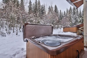 Un jacuzzi exterior con revestimiento de madera, rodeado de árboles cubiertos de nieve y una terraza de madera.