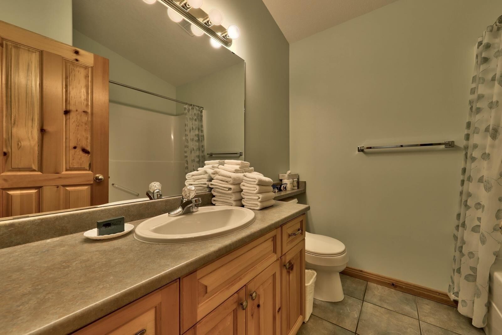 A bathroom with a wooden vanity, a sink, a mirror with lights, a toilet, and a shower curtain.