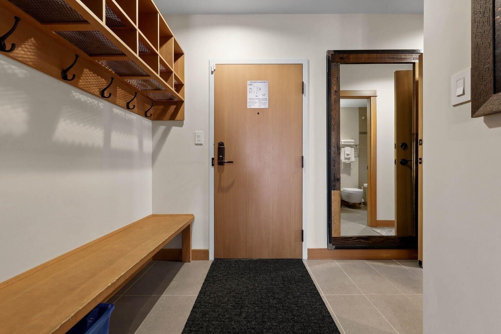 A hallway with a wooden bench, a door with a notice, and a mirror reflecting a bathroom.