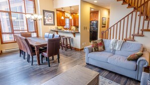 A spacious living room with a grey sofa, wooden dining table, and chairs. A kitchen with wooden cabinets and a refrigerator is visible in the background. - Woodhaven #32 By Bear Country (Sun Peaks)