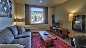 Una sala moderna con un sofá, una mesa de centro, un televisor sobre un mueble de madera y un ventanal con vistas a árboles.