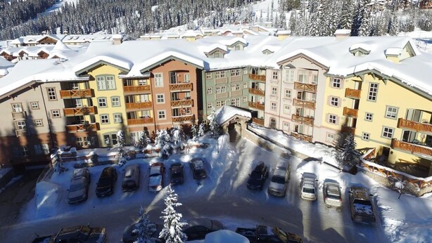 A multi-story hotel with balconies, a parking lot, and snow-covered surroundings. - Fireside Lodge #413 By Bear Country (Sun Peaks)