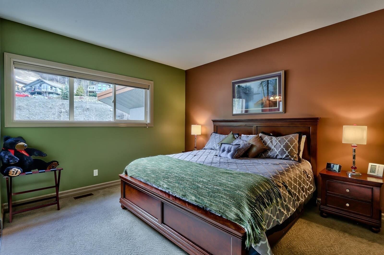 A bedroom with a large bed, two bedside tables, a window, and a teddy bear on a small table.