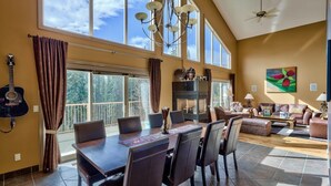A spacious living room with a high ceiling, large windows, a fireplace, and a dining area with a table and chairs.