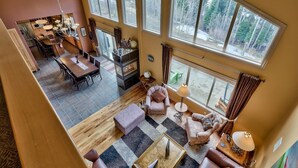 A spacious living area with a dining table, sofas, and a fireplace. Large windows offer views of a forested area.