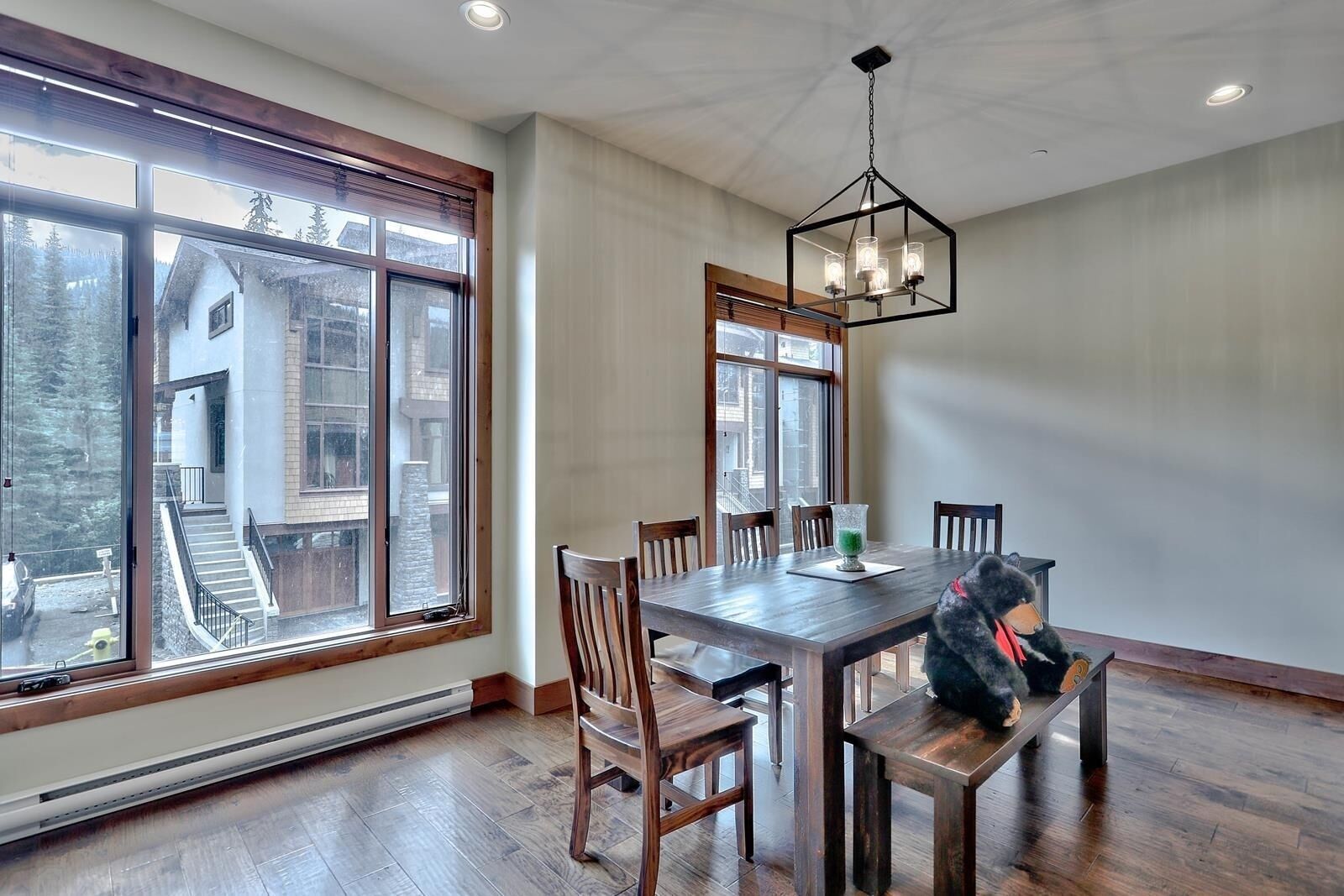Comedor con mesa y sillas de madera, ventanas amplias con vistas a un edificio y árboles, y una lámpara colgante.