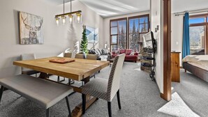 Una sala de estar moderna con una mesa de comedor de madera, un sofá y una televisión. Hay un ventanal con persianas, un pequeño árbol de Navidad y una chimenea.