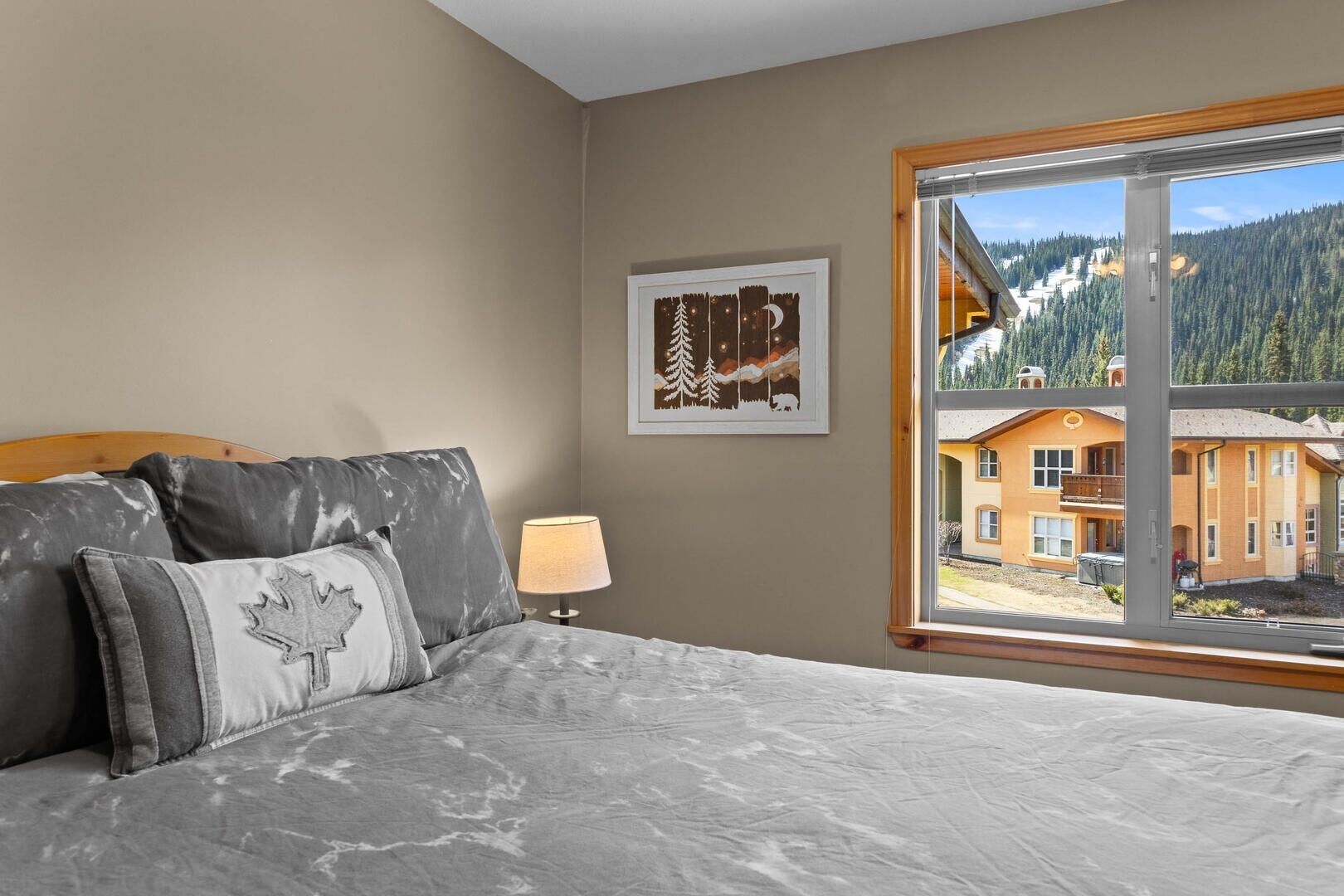 A bedroom with a bed, a nightstand, a lamp, and a window with a view of a mountain lodge.