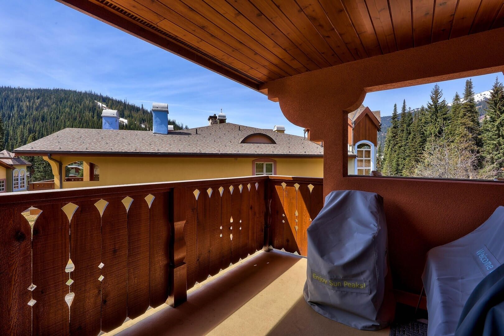 A balcony with a wooden railing, a covered grill, and a view of a mountain and a house.