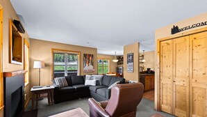 A cozy living room with a fireplace, a leather armchair, a sofa, and a wooden table. There is a large wooden door with a 'WELCOME' sign above it. - Crystal Forest #22 By Bear Country (Sun Peaks)