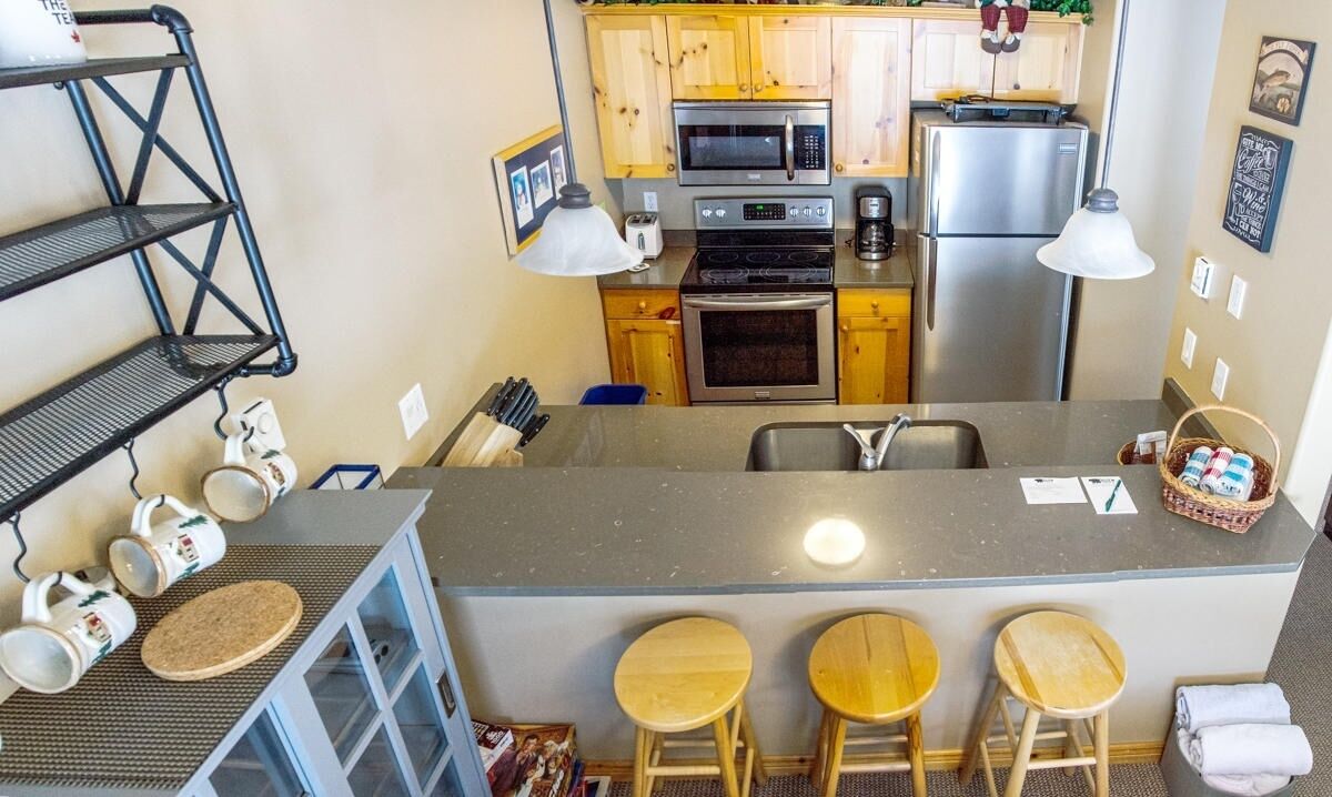 A compact kitchen with a central island, wooden cabinets, a microwave, oven, and a refrigerator. There are bar stools and a coffee machine.