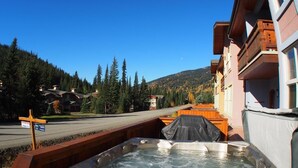 A hot tub in a wooden deck area with a mountain view, surrounded by buildings and trees.