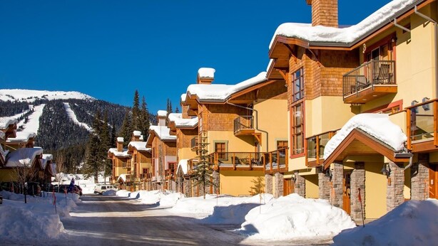 A row of log cabin-style buildings with snow-covered roofs, set against a backdrop of a clear blue sky and a snow-covered mountain. - Trails Edge #25 By Bear Country (Sun Peaks)
