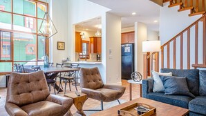 A spacious living room with leather armchairs, a blue sofa, and a dining area with a table and chairs. A kitchen with wooden cabinets and a refrigerator is visible in the background. - Trails Edge #25 By Bear Country (Sun Peaks)