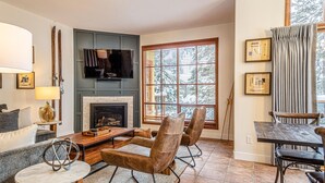 A living room with a fireplace, a television mounted above it, and a dining area with chairs and a table. - Trails Edge #25 By Bear Country (Sun Peaks)