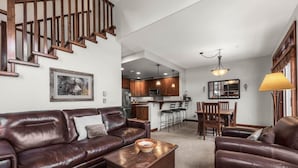 A spacious living room with a brown leather sofa, a wooden coffee table, and a dining area with wooden chairs and a table. A kitchen with wooden cabinets and a refrigerator is visible in the background. - Trappers Landing #11 (Sun Peaks)