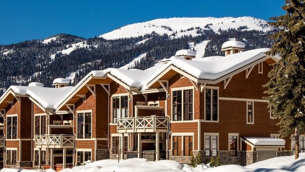 A row of wooden cabins with snow-covered roofs, set against a backdrop of snow-covered mountains and clear blue sky. - Stones Throw #56 By Bear Country (Sun Peaks)