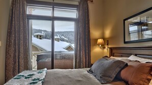 A hotel room with a large bed, a view of snow-covered buildings, and a window with curtains. - Stones Throw #56 By Bear Country (Sun Peaks)