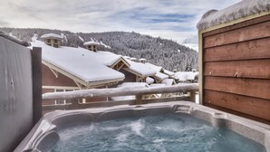 A hot tub with a wooden deck, overlooking a snowy mountain landscape and several buildings. - Stones Throw #56 By Bear Country (Sun Peaks)