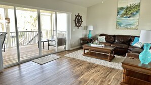 Ein geräumiges Wohnzimmer mit Holzboden, einem braunen Ledersofa, einem Couchtisch und einer Glastür, die auf eine Terrasse führt.