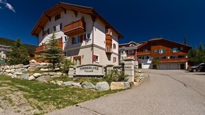 A multi-story building with a stone facade, a sign reading "Timberline Village," and a parking area with a stone wall and greenery. - Timberline Village #38 By Bear Country (Sun Peaks)