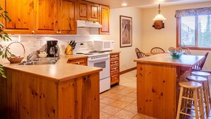 A kitchen with wooden cabinets, a stove, a microwave, and a sink. There is a dining area with a table and chairs. - Timberline Village #38 By Bear Country (Sun Peaks)