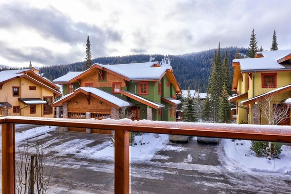 A row of colorful wooden cabins with snow-covered roofs, set against a backdrop of evergreen trees and a cloudy sky.
