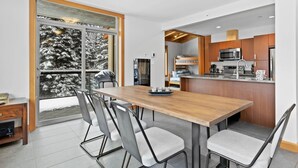 A modern kitchen with a wooden dining table, grey chairs, and a view of snow-covered trees through a glass door.