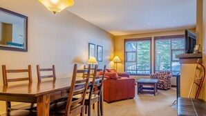 A living room with a wooden dining table, chairs, a sofa, and a TV. There are windows with blinds and a lamp on the table. - Stones Throw #11 By Bear Country (Sun Peaks)