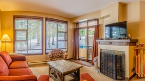 A cozy living room with a fireplace, a sofa, a chair, and large windows. - Stones Throw #11 By Bear Country (Sun Peaks)