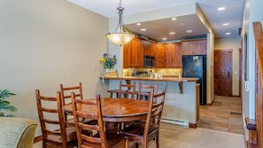 A dining area with a wooden table and chairs, a kitchen with wooden cabinets and a refrigerator, and a hallway with wooden doors. - Trappers Landing #06 By Bear Country (Sun Peaks)