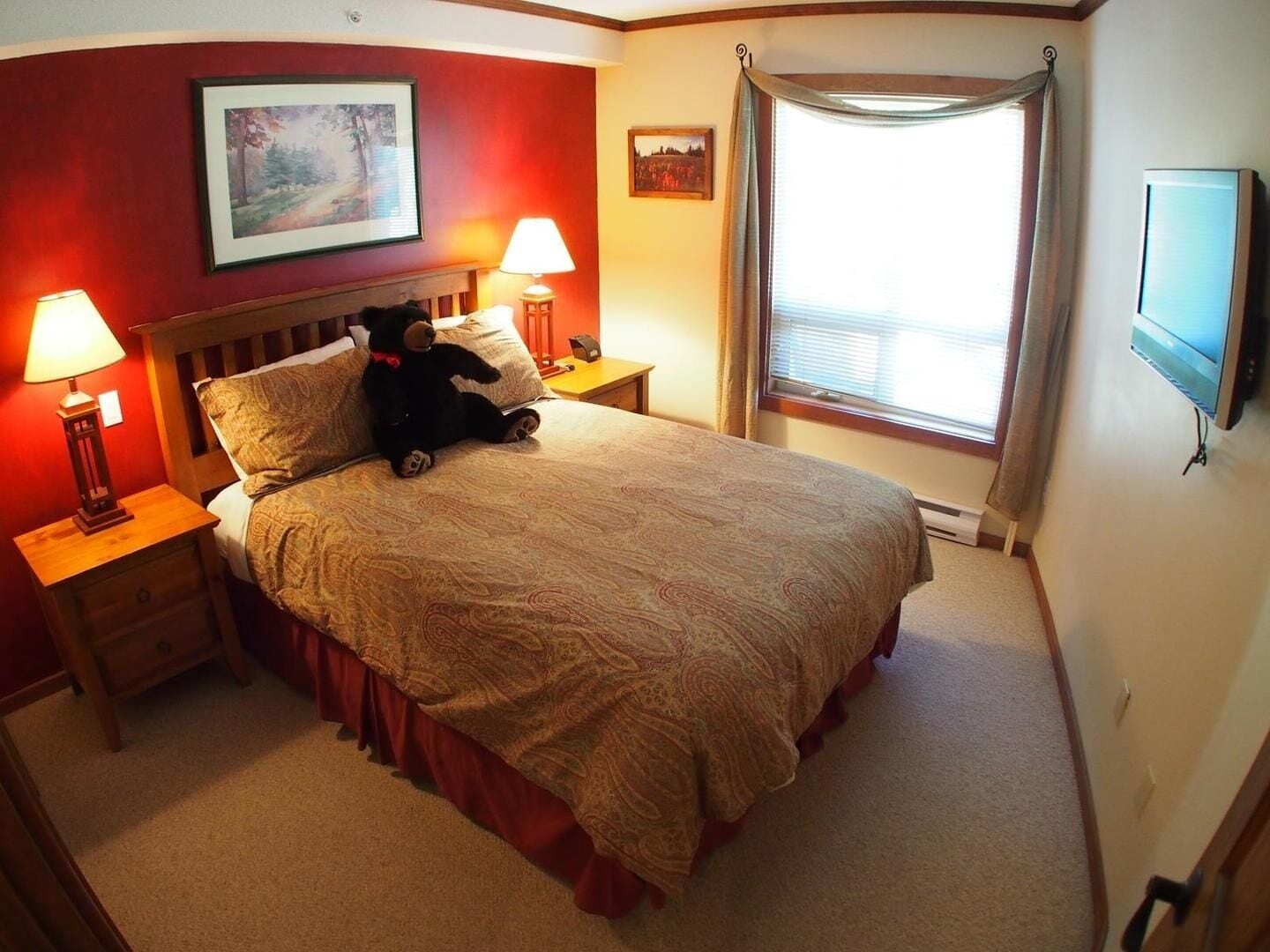 A neatly made bed with a patterned bedspread, two bedside lamps, a teddy bear, and a painting on the wall.