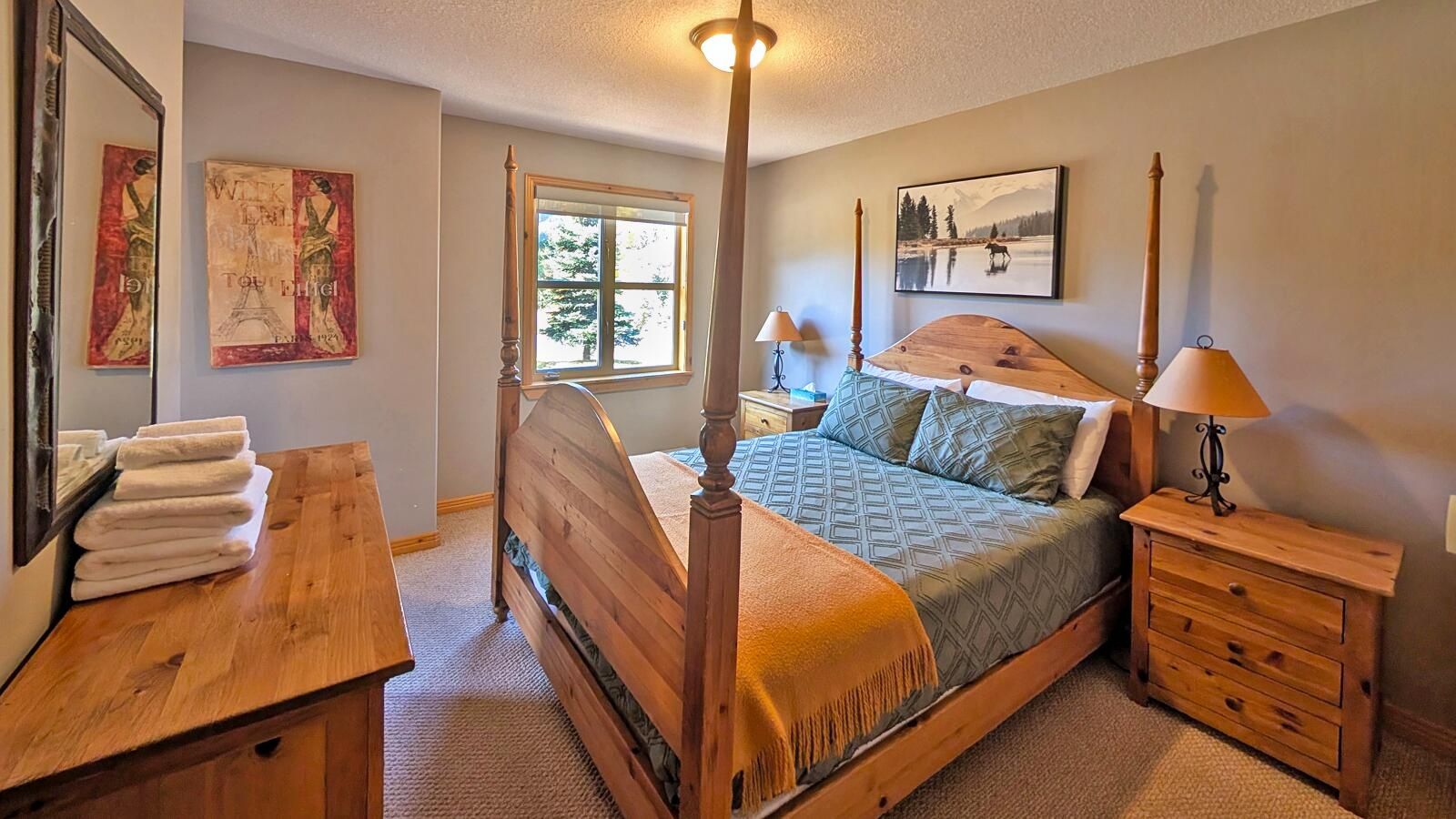 A bedroom with a four-poster bed, a wooden dresser, a nightstand, and a window with a view of trees.