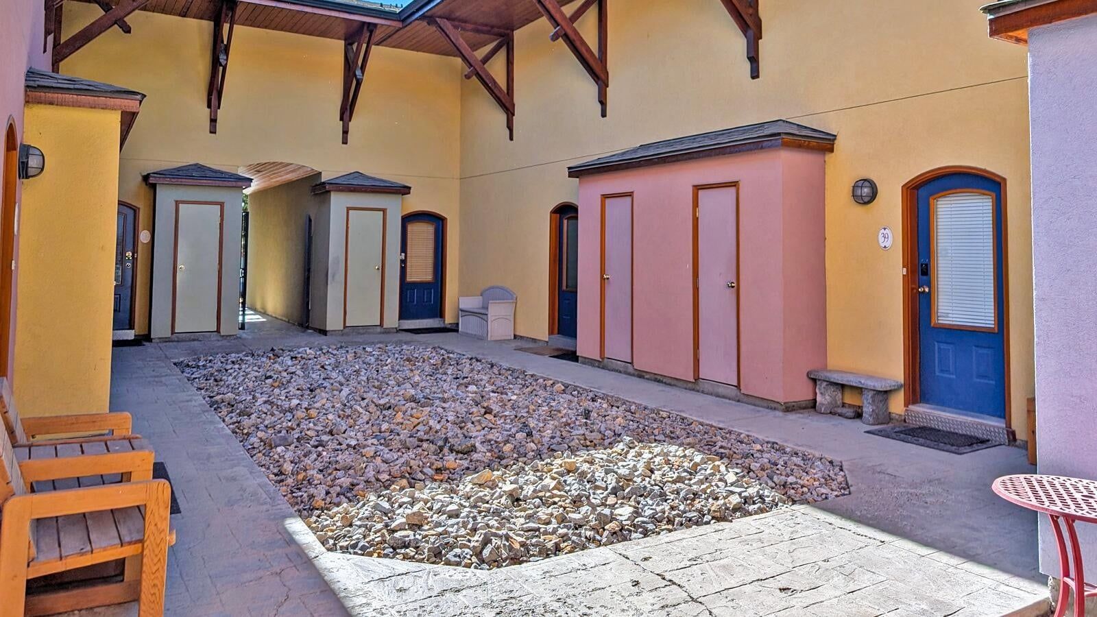 A courtyard with a stone floor, wooden benches, and a building with multiple doors and windows.